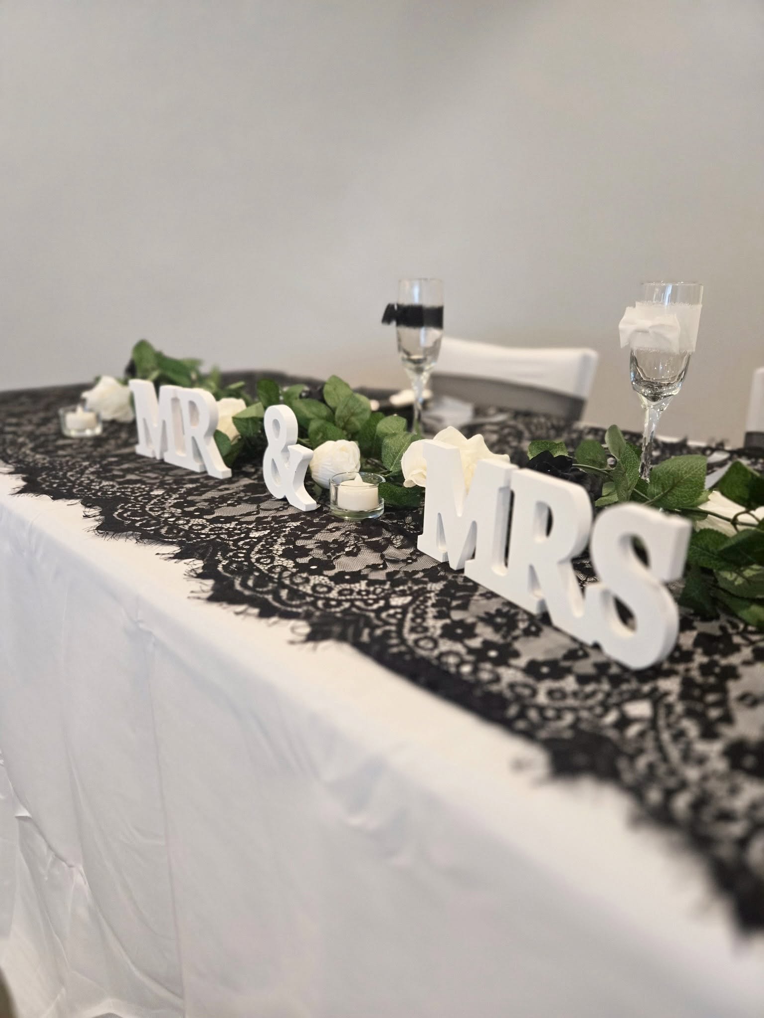 Table with black lace runner, greenery, white roses, candles and champagne flutes
